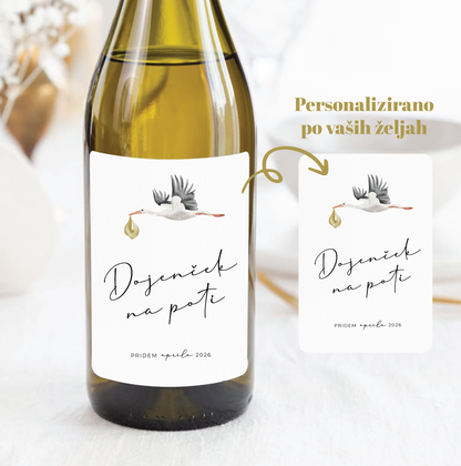 Personalizirana vinska etiketa s štorkljo in napisom »Dojenček na poti« na steklenici belega vina.